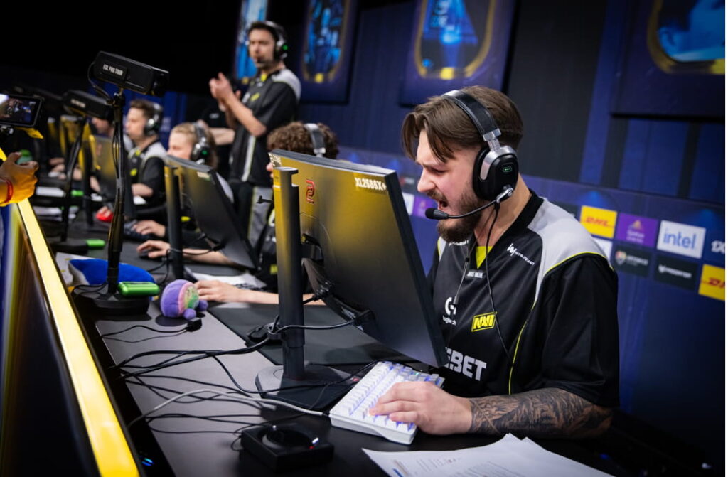 En e-sportspelare i IEM Katowice med lagkamrater i bakgrunden vid datorer på en turneringsscen.