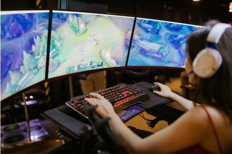 Kvinna spelar datorspel med headset framför tre skärmar i en gamingmiljö.