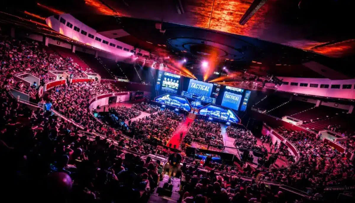 Fullsatt arena med publik som tittar på en e-sportturnering med färgglada ljus och stora skärmar på scenen.