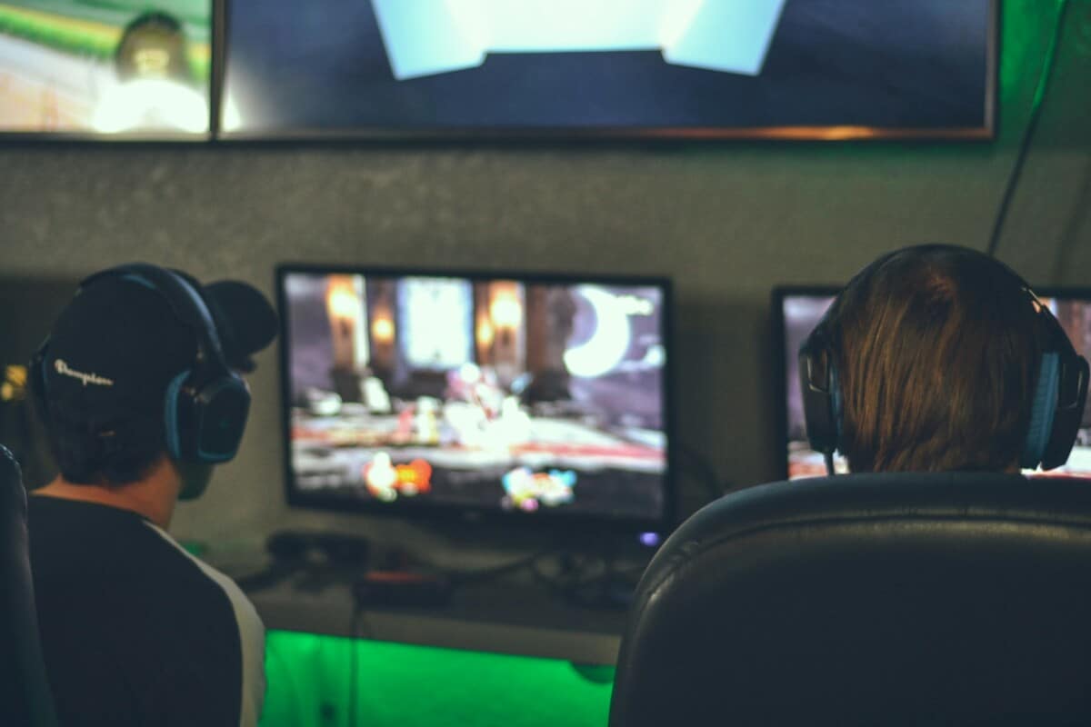 Två personer med headset spelar tv-spel framför skärmar i ett mörkt rum.
