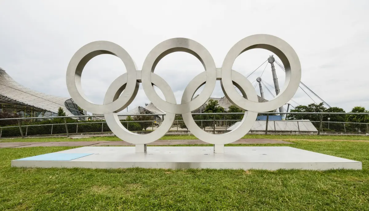 Stora vita olympiska ringar som konstverk utomhus, med gräs och byggnader i bakgrunden.