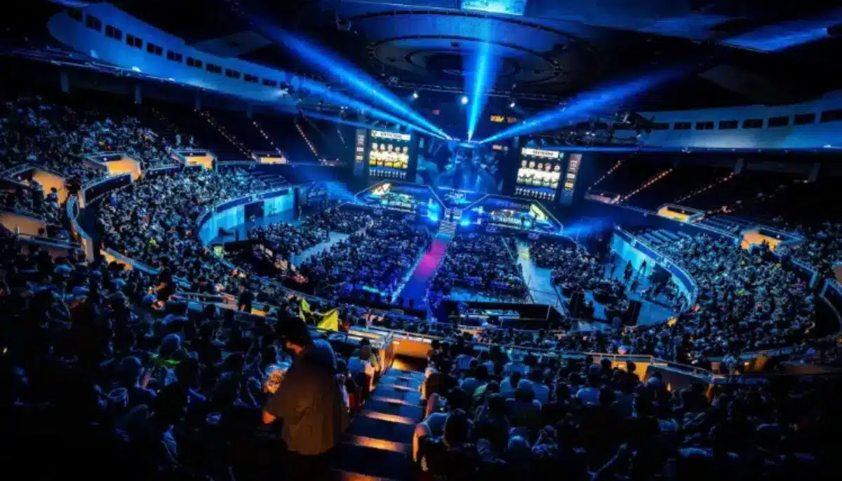 Stor e-sportarena fylld med publik, scen, strålkastare och stora skärmar som visar matchen.