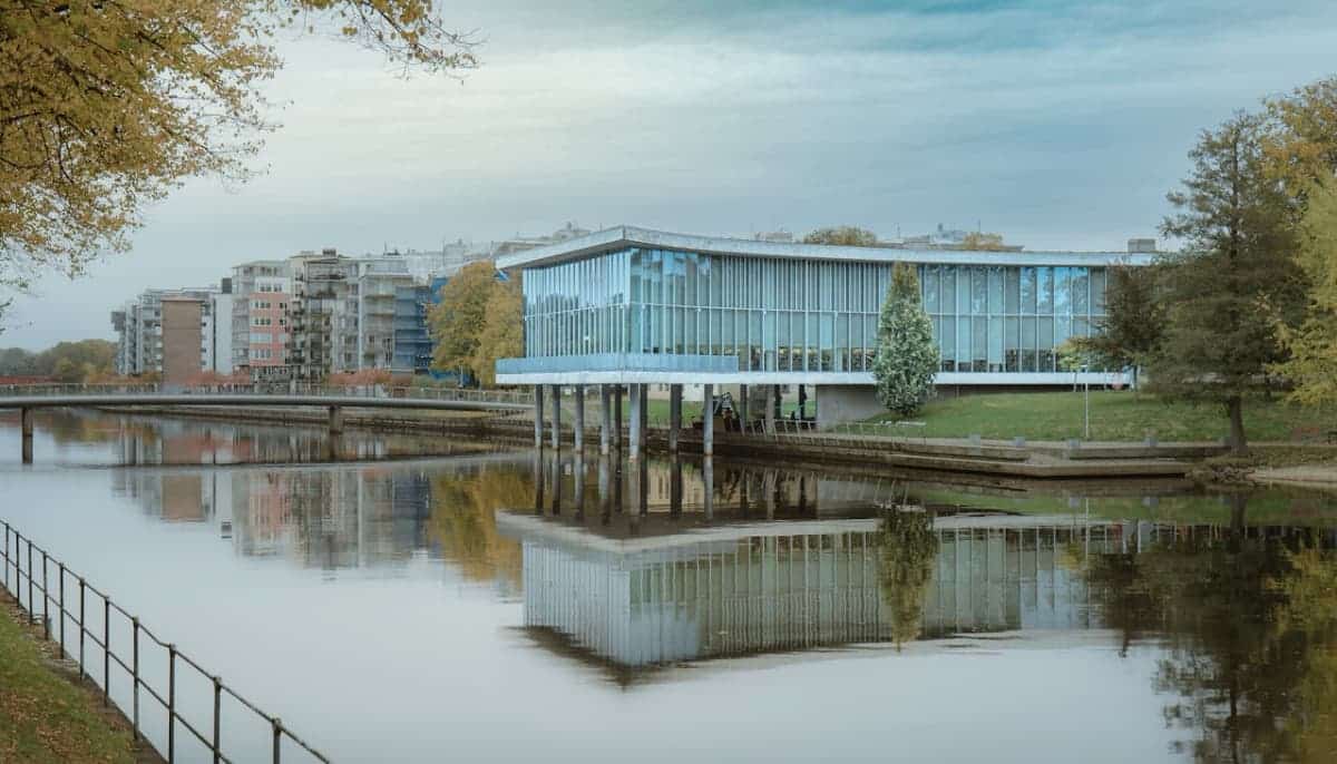 Modern byggnad med glasfasad vid vattnet i stadsmiljö, speglande i den lugna ytan.