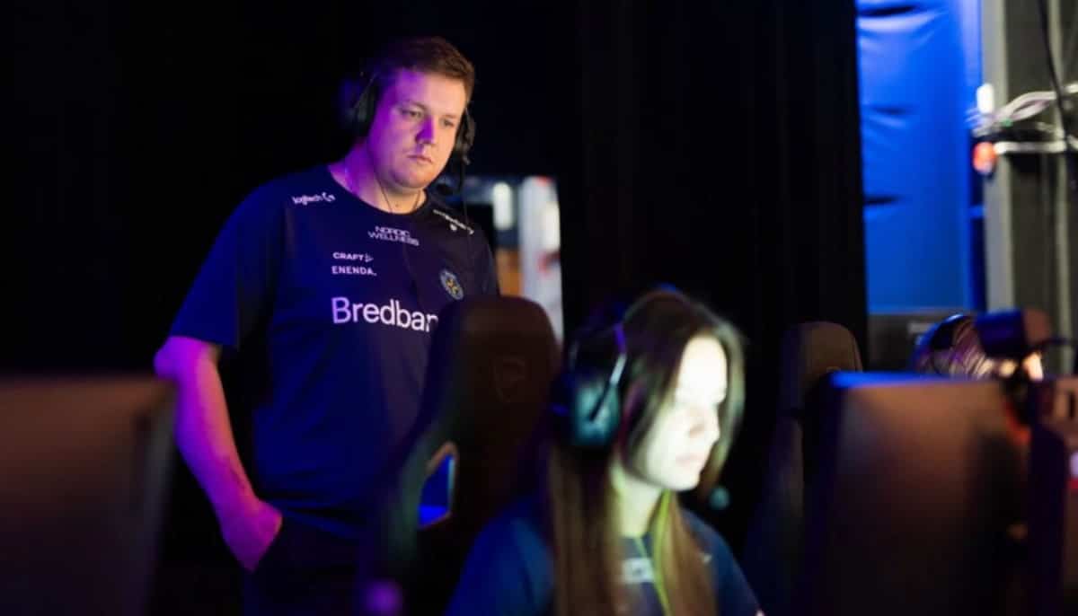 Coach med headset står bakom e-sportspelare vid datorer under match i upplyst spelmiljö