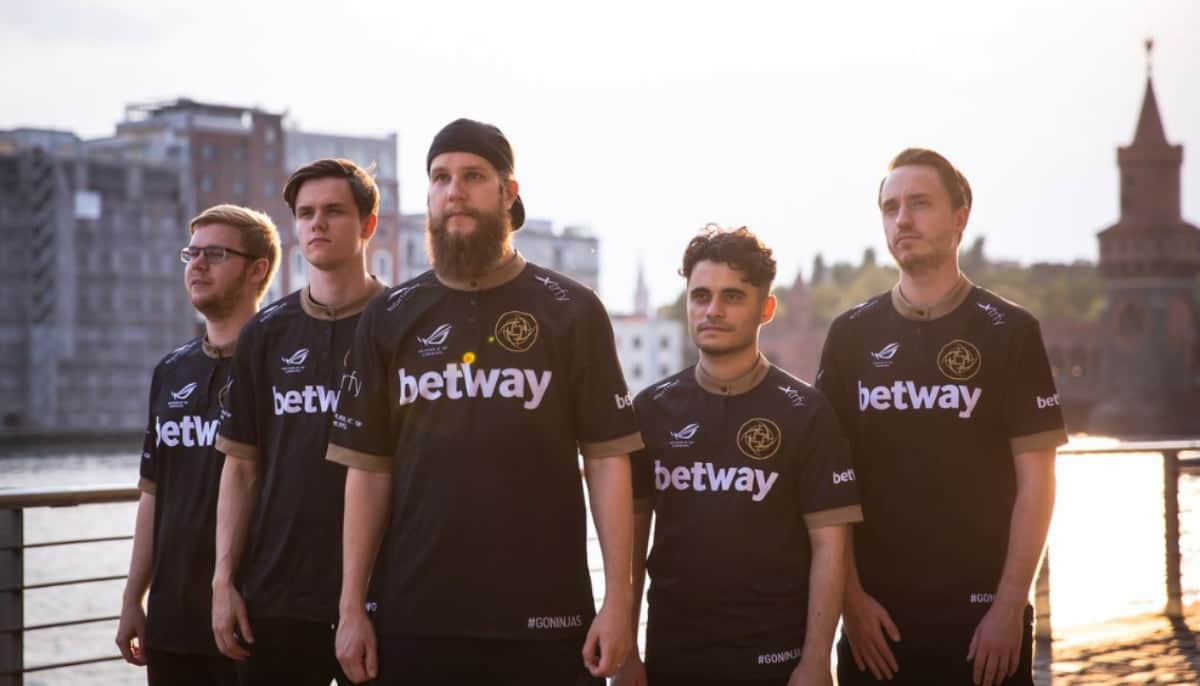 E-sportlag i svarta lagtröjor poserar utomhus vid vattnet med stadsmiljö i bakgrunden i solnedgång.