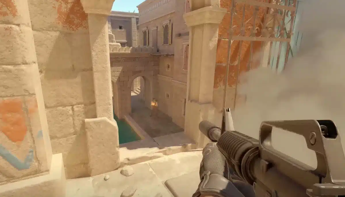 Förstapersonsvy från Counter-Strike 2 där spelare siktar med gevär i sandfärgad bana med rök och explosion i bakgrunden.
