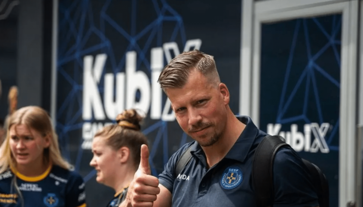 En man i sporttröja ger tummen upp framför en Kublix-bakgrund, med två personer i lagkläder bakom honom.