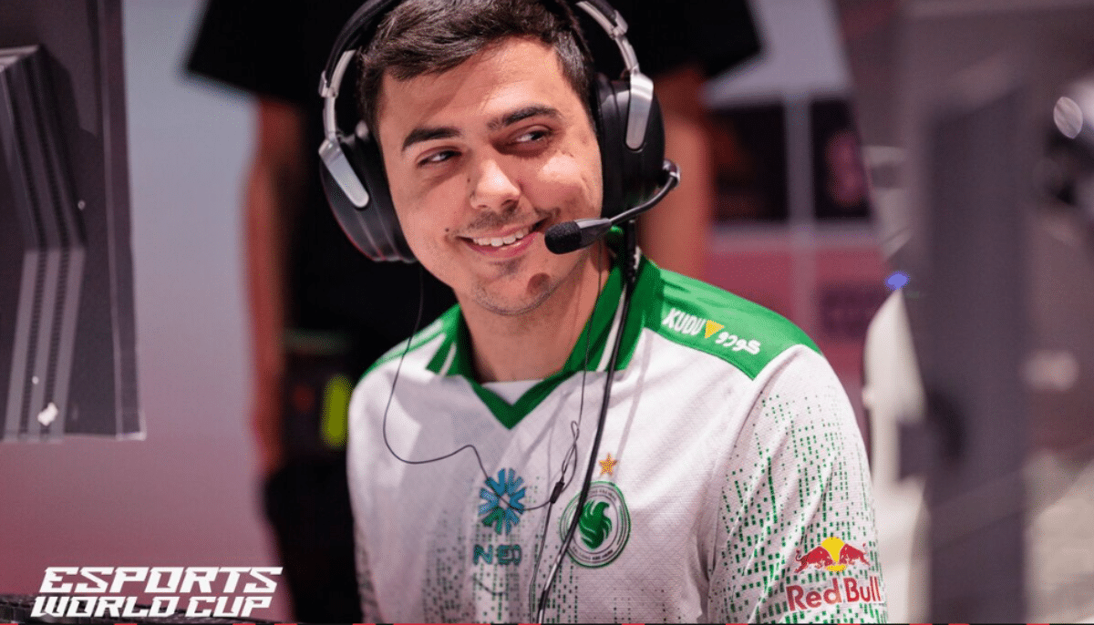 E-sportspelare med headset ler vid dator under Esports World Cup, iklädd vit och grön matchtröja.