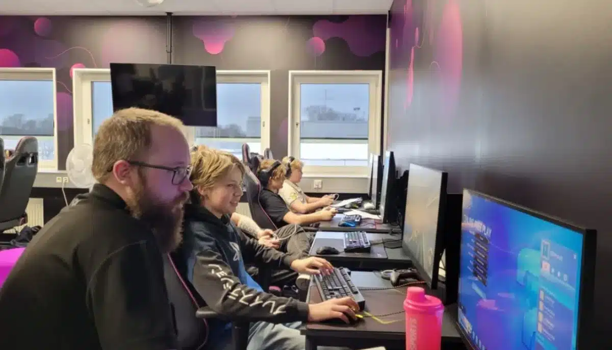 Deltagare sitter vid datorer och tränar e-sport i ett klassrum med gamingstolar, skärmar och instruktör närvarande.