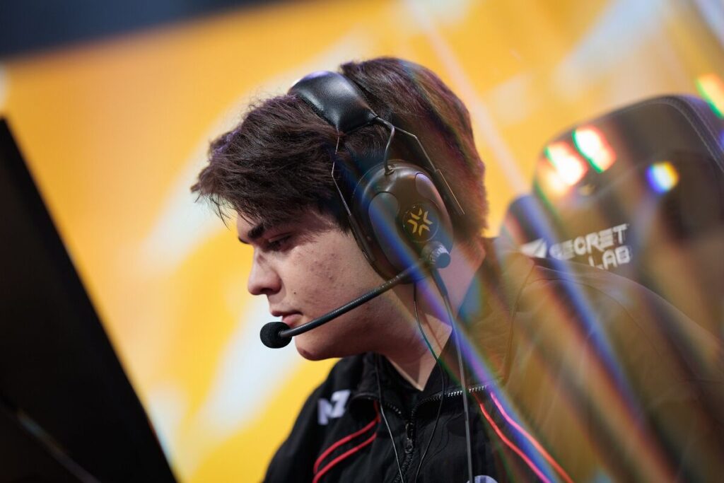 Evv wearing a headset, seated in front of a monitor, immersed in an esports competition, with a vibrant background.