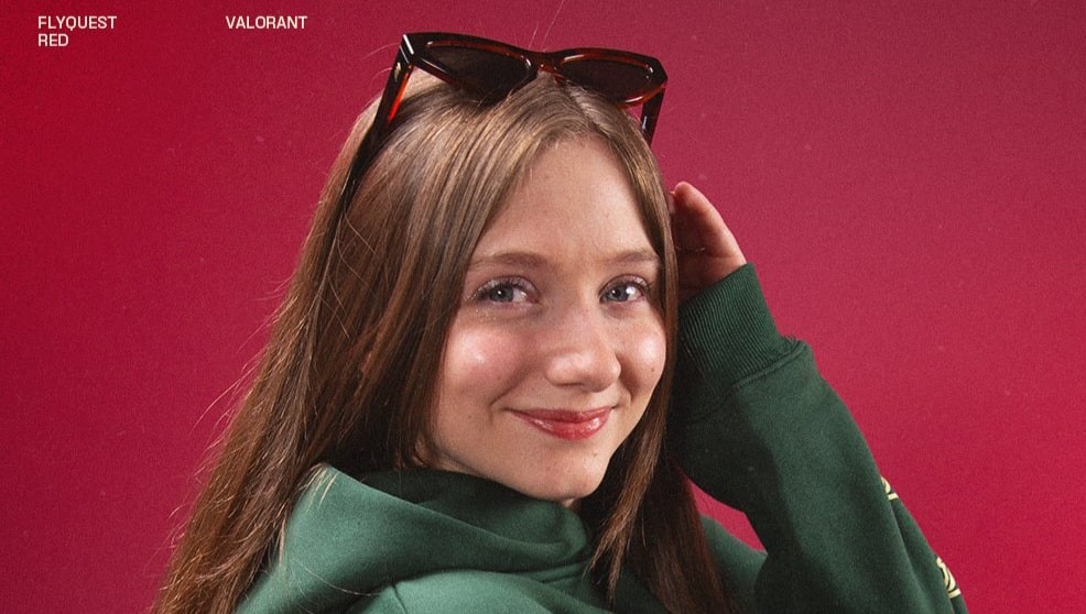 Theia wearing a green hoodie and sunglasses poses against a vibrant red background, showcasing a stylish, casual look.