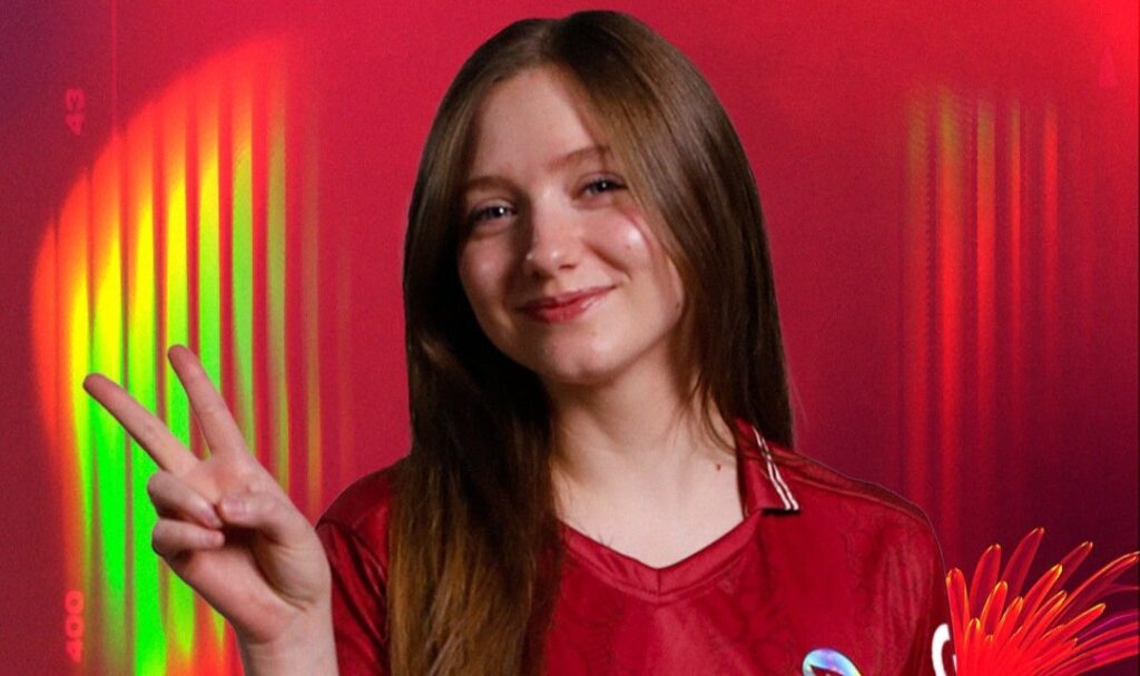 Theia in a red jersey poses against a vibrant red and green background, flashing a peace sign with their hand.