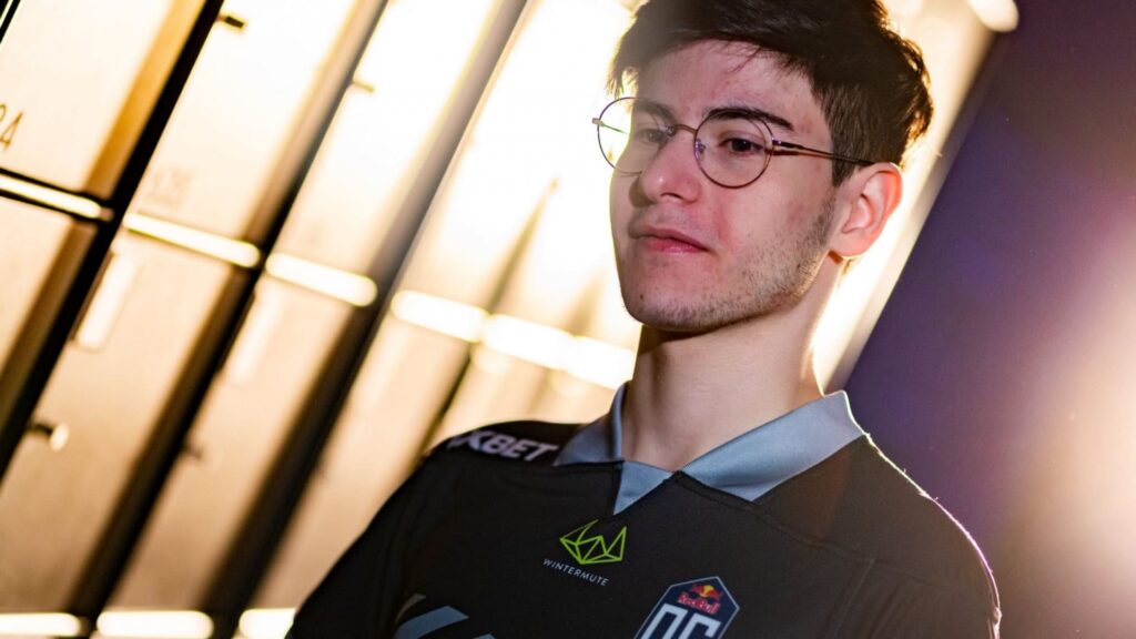 Shahar ‘FlameZ’ Shushan wearing a black esports jersey stands in front of illuminated lockers, with a blurred face and glasses in a dynamic pose.