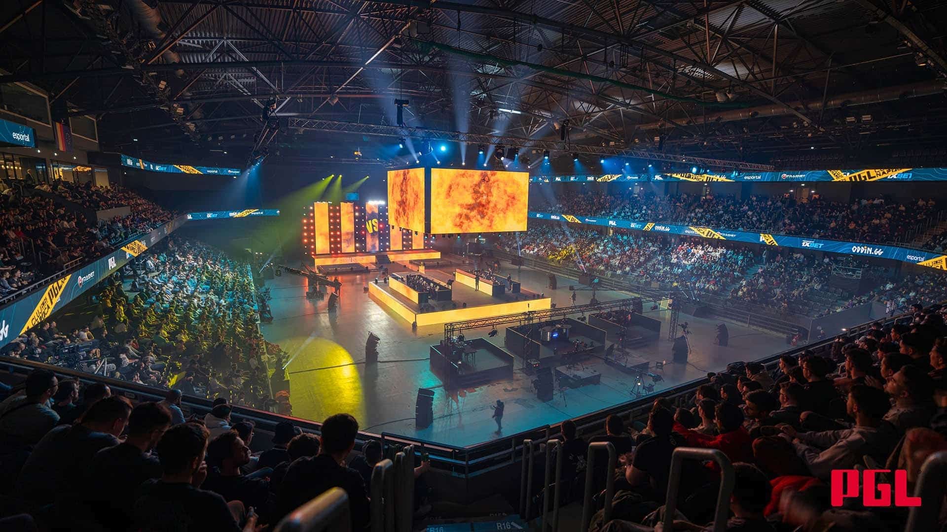 A large, packed sports arena during an esports tournament, showing thousands of spectators watching players on stage under bright lights and a large central video screen