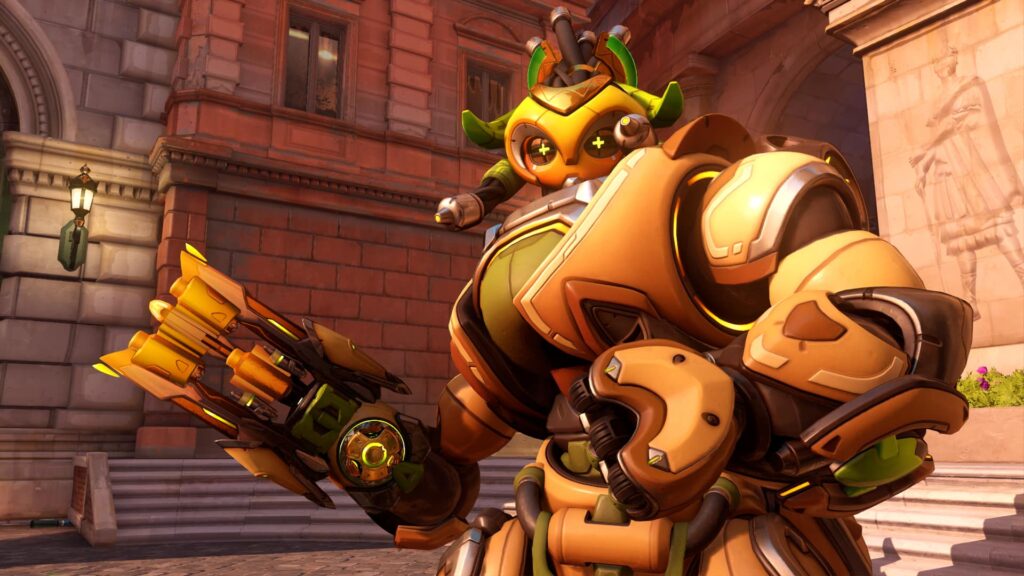 Orisa from Overwatch in golden armor aiming her weapon, standing in a city courtyard with red brick and stone buildings in the background.