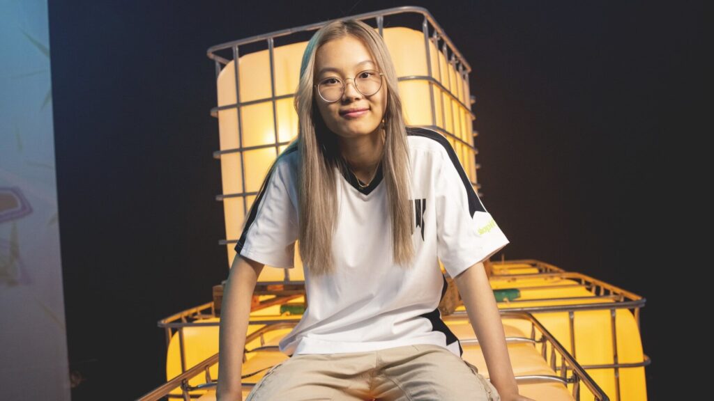 Sonder, a young female esports player wearing glasses and a white team jersey, sits on a lit stage setup