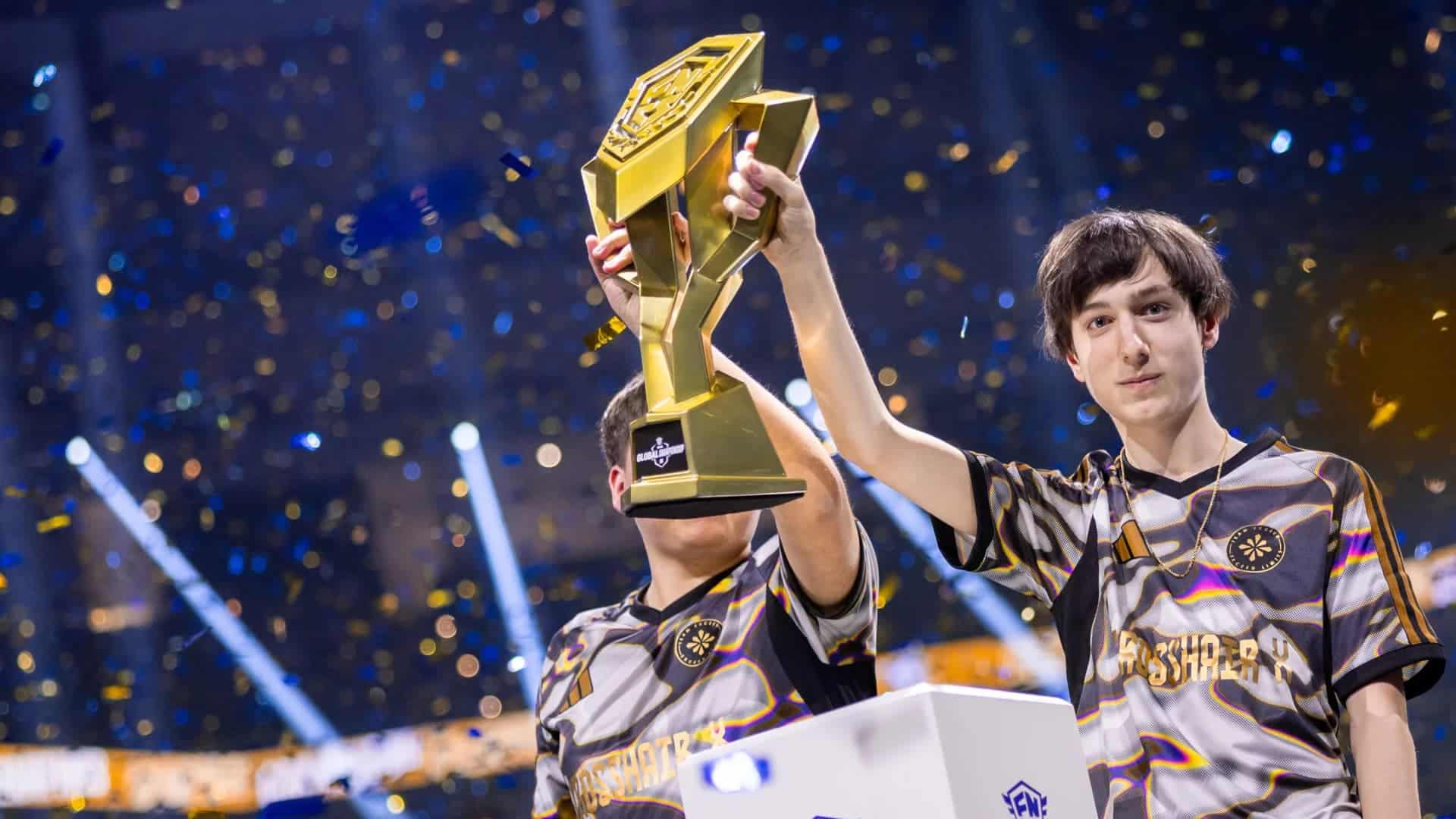 Two young men in esports jerseys hold up a large golden trophy in celebration