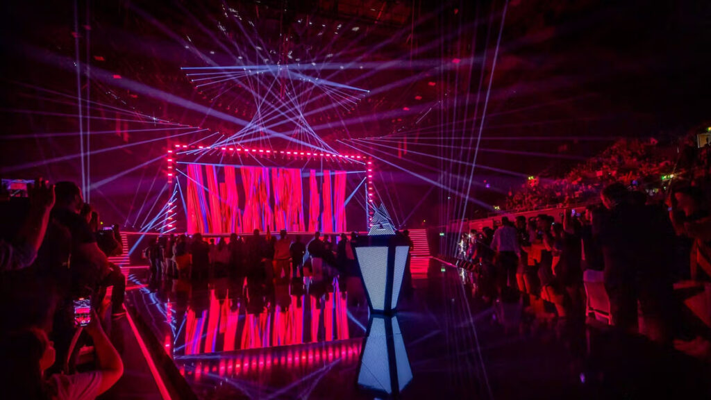 Image of BLAST Premier trophy on a platform. A stage lit up with blue and red lighting is illuminated in the background with a crowd of esports fans sitting and standing in front of it.