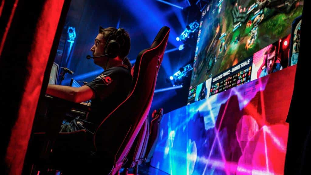 Pro esports player at a tournament stage, with headset on and a large screen showing a League of Legends match.