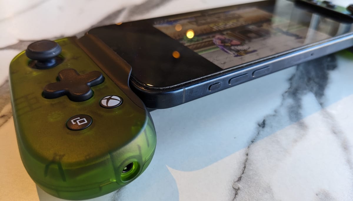 A green handheld game controller attached to a smartphone displaying a fighting game on its screen, set on a marble surface