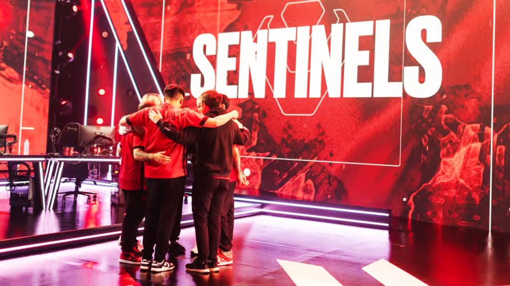 Image of Sentinels huddling together on an esports event stage. The Sentinels name and logo is projected on the screen in the background