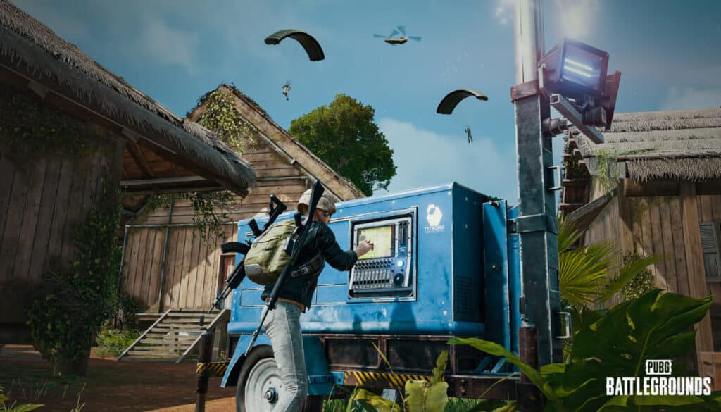 A player from PUBG interacts with a blue supply drop machine while paratroopers descend nearby, framed by rustic wooden buildings and greenery.