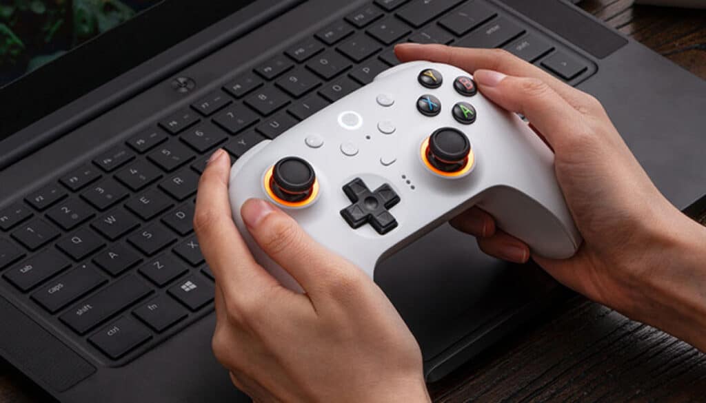 Person holding a white 8BitDo Ultimate 2 controller with illuminated thumbsticks in front of a laptop