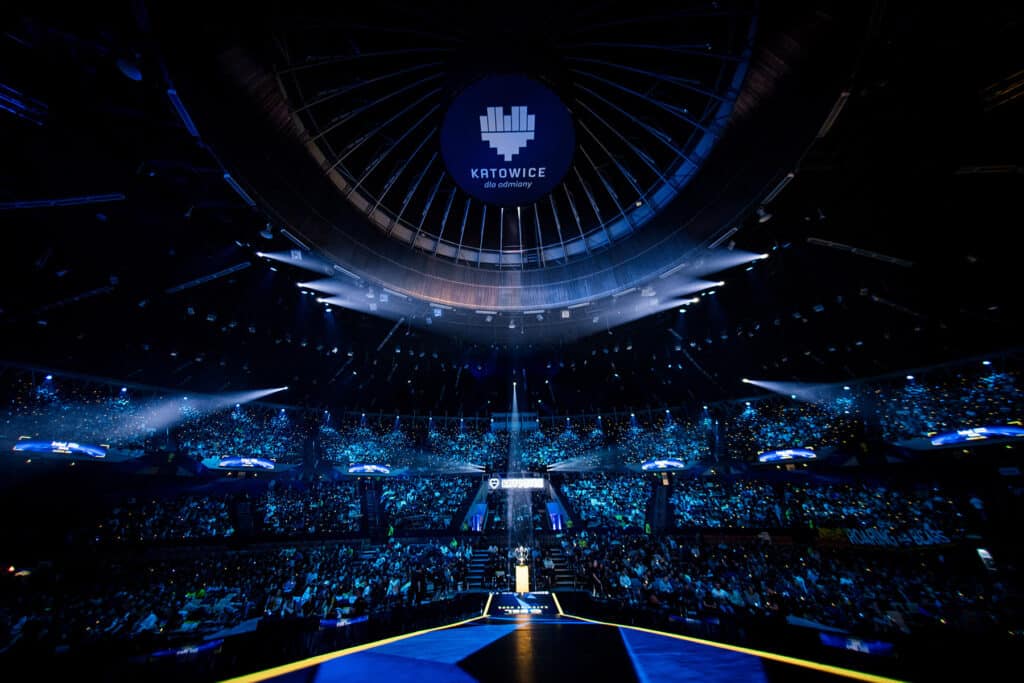 Image of ESL One Katowice stage lit up with stage lighting. The Katowice heart logo is on the ceiling of the venue with a light illuminating a trophy on the stage