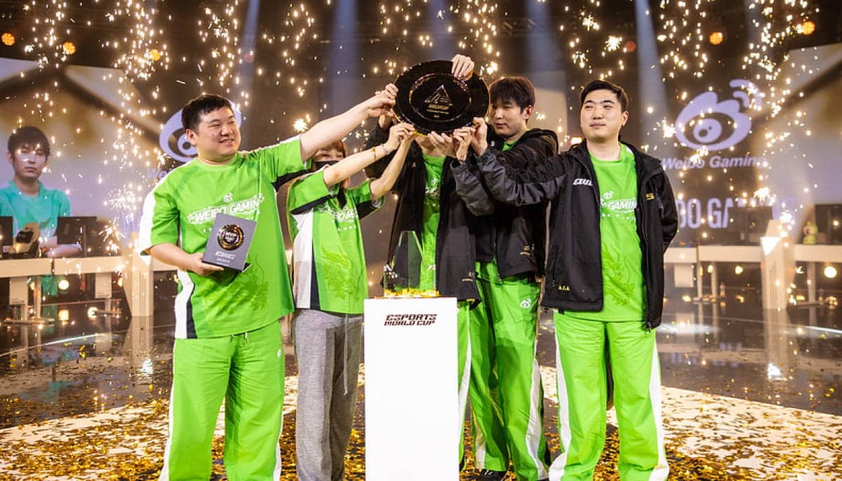 Five esports team members wearing green uniforms celebrate a victory. They hold up a trophy amidst sparkling confetti and bright stage lights, exuding joy and triumph