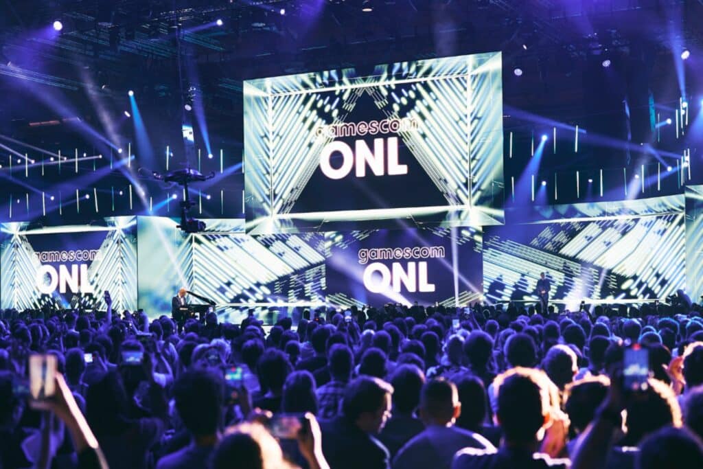 Image of Gamescom Opening Night Live stage illuminated with lights and branding. A crowd of people is looking at the stage in the foreground