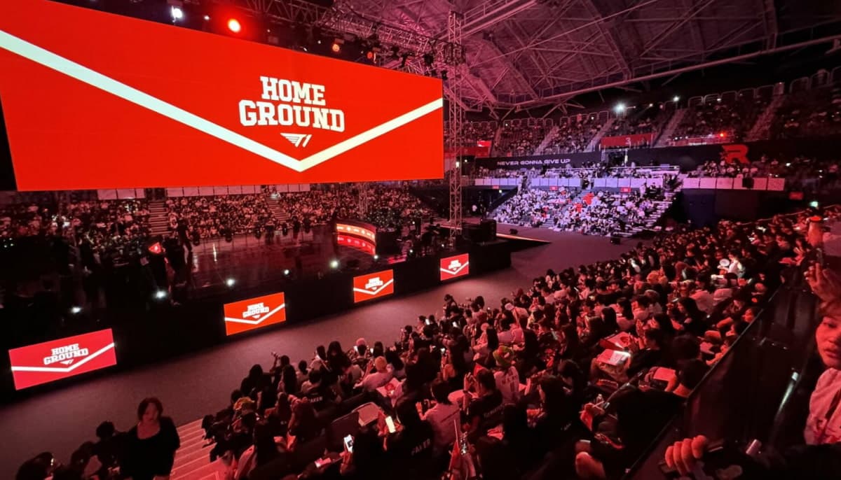 A large indoor arena filled with people watching a bright red screen displaying “Home Ground”
