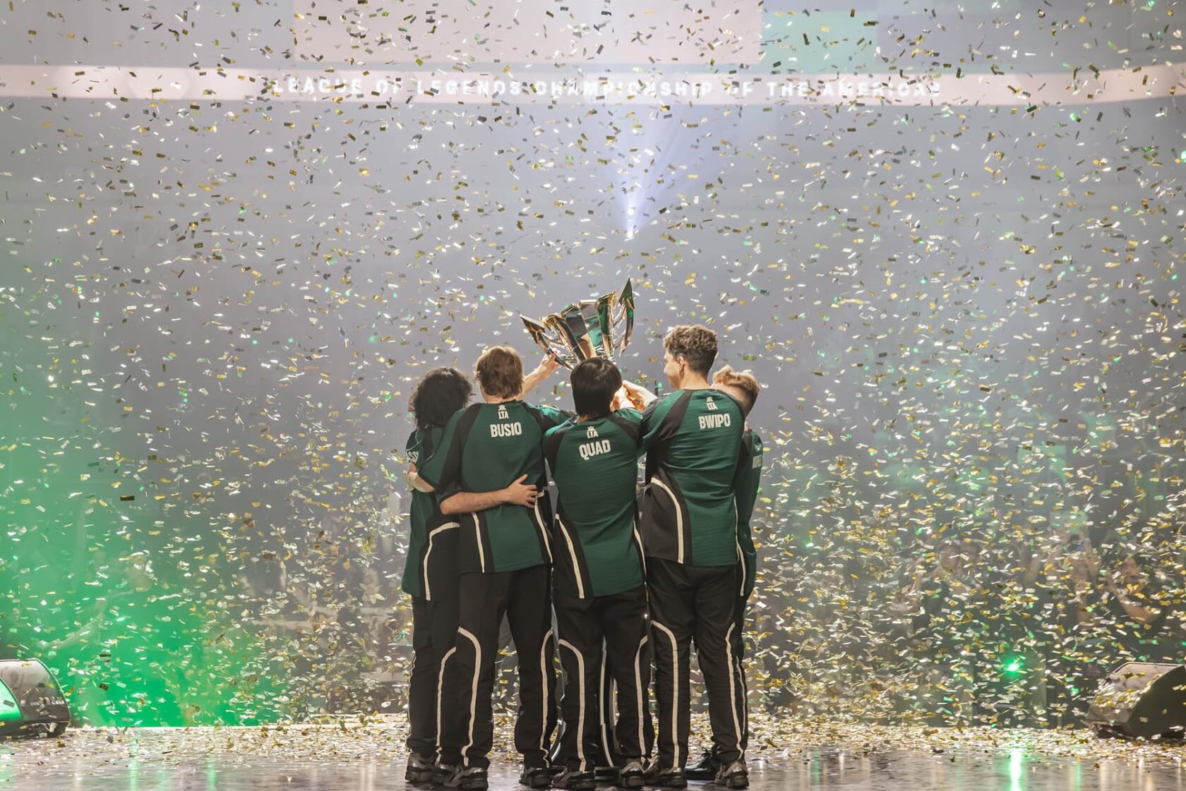 Image of FlyQuest lifting LTA North trophy on a stage. Confetti is covering the stage and the crowd in the background