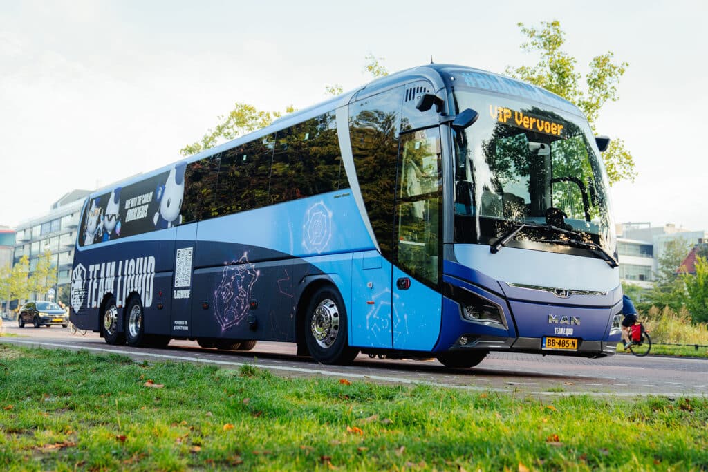 Image of a blue Team Liquid-branded bus parked on a road. A patch of grass is in the foreground