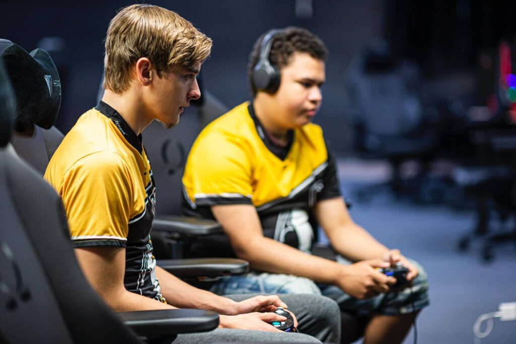 Two players in yellow and black jerseys playing in an esports facility with controllers in hand