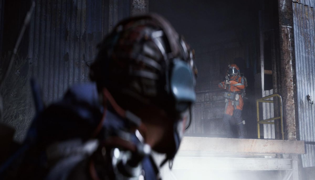 A person in a dark helmet and gear stands in the foreground, looking towards another person in an orange suit and helmet inside a dim, industrial building in Arc Raiders
