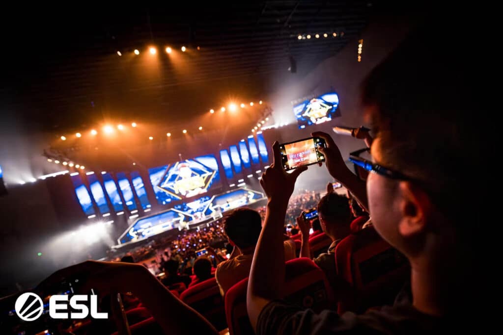 Image of IEM Chengdu main stage illuminated with warm lighting. A fan in the audience is taking a picture of the stage on their phone