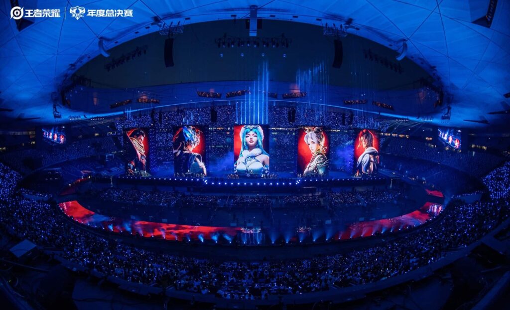 Wide-angle view of an esports arena with vibrant blue lighting, large screens displaying colorful characters, and an audience immersed in the event