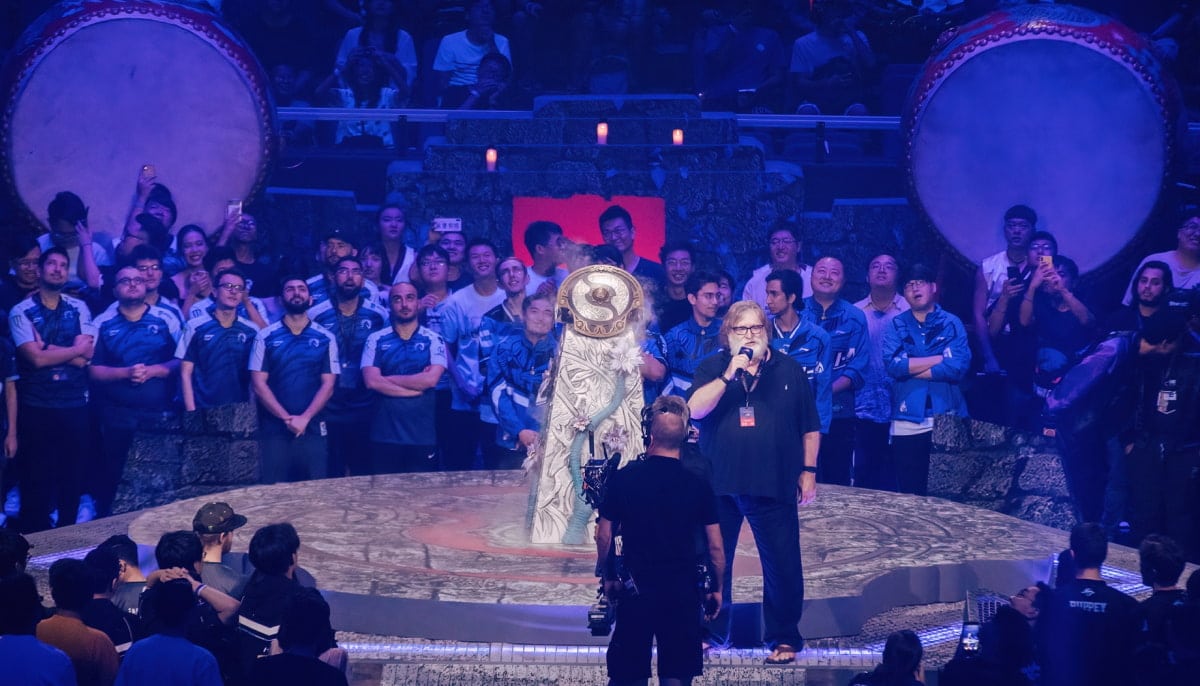 A stage view of the opening ceremony for The International 2019 Dota 2 championship shows Gabe Newell of Valve speaking into a microphone, flanked by two large ceremonial drums and players from the competing teams wearing blue and black team jerseys on an illuminated stage