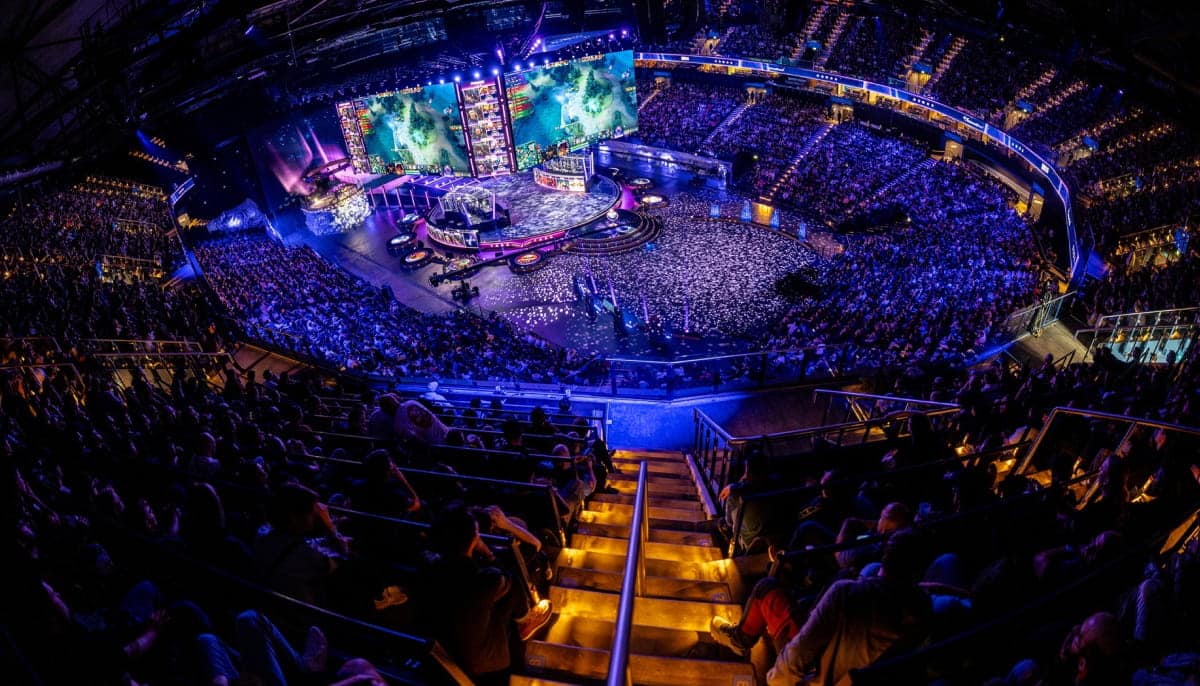 A wide-angle shot of a large, dark arena at The International (TI) esports event, filled with thousands of spectators watching a Dota 2 match on massive screens above a central stage