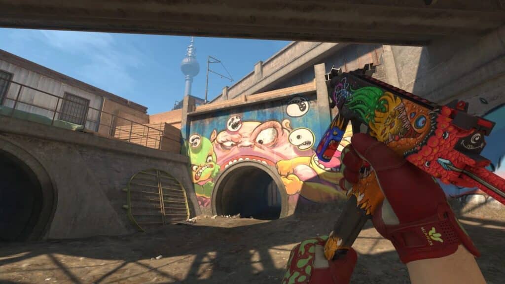 A first-person view from the video game Counter-Strike 2 shows a player holding an MP9 submachine gun with a colourful "Food Chain" skin and red "Crimson Web" gloves, in front of a tunnel with a large monster graffiti mural on the Berlin map
