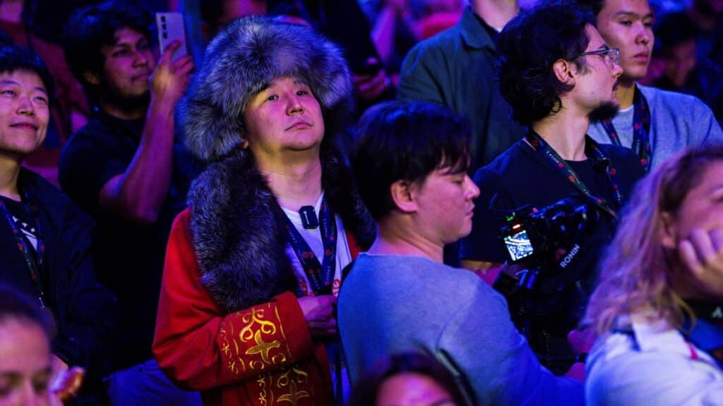 A man in a large fur hat and a red, embroidered robe is seated in a crowd of spectators, looking up at an event stage. Other people around him wear casual clothing and event lanyards. One person nearby is operating camera equipment labeled "RONIN"