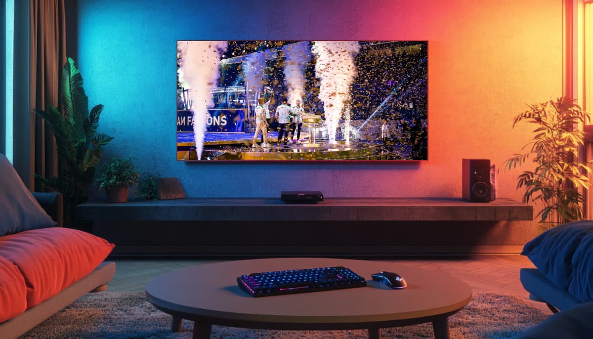 The image shows a living room with a TV broadcasting an esports event. The screen shows players from the Team Falcons esports team celebrating their victory