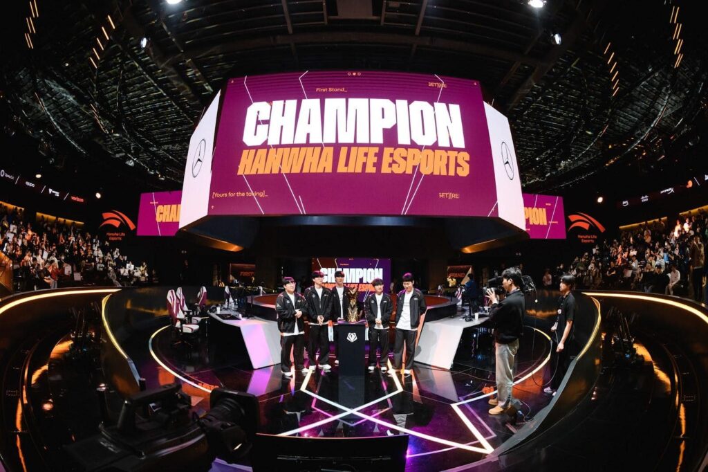 Five professional eSports players stand together on a stage holding a trophy and an LCK pennant as confetti rains down. A large screen behind them displays the words "CHAMPION HANWHA LIFE ESPORTS"