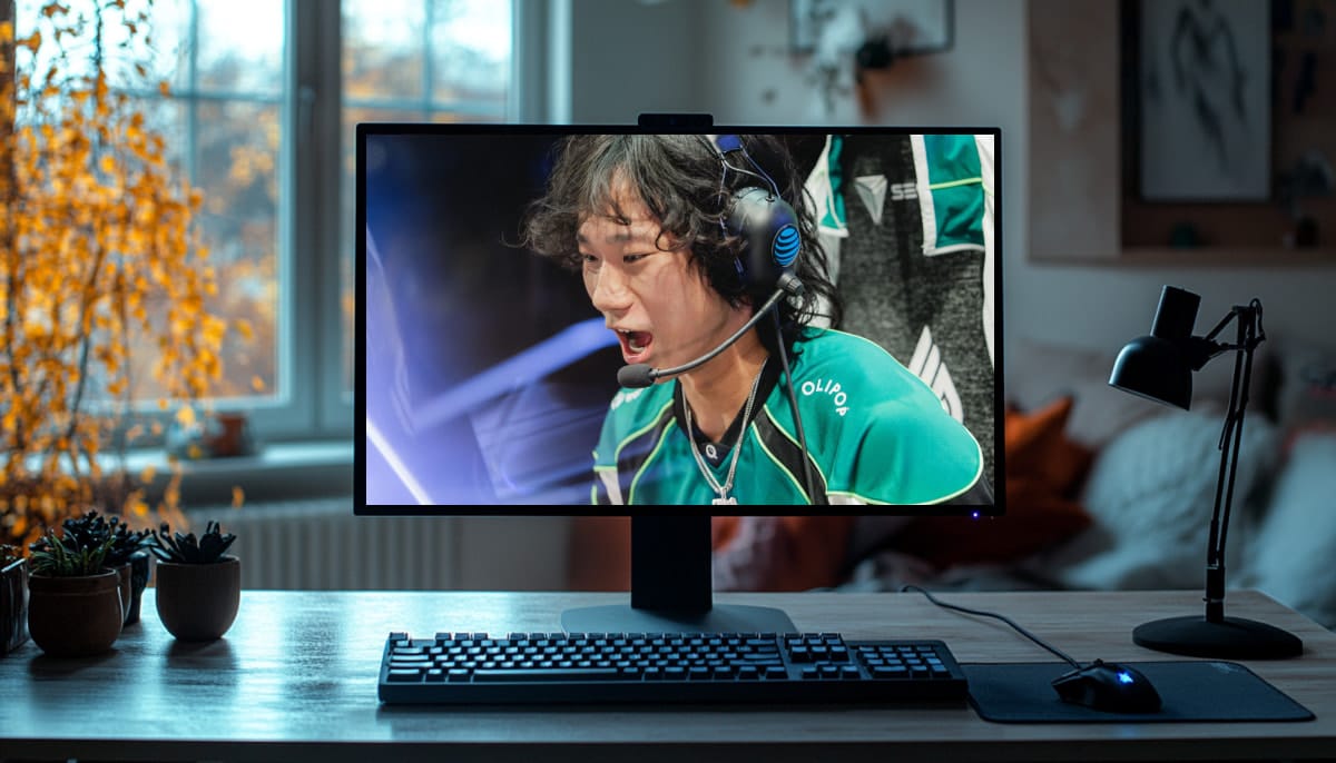 A young male esports player wearing a green jersey and a gaming headset shouts intensely during a match shown on a computer monitor