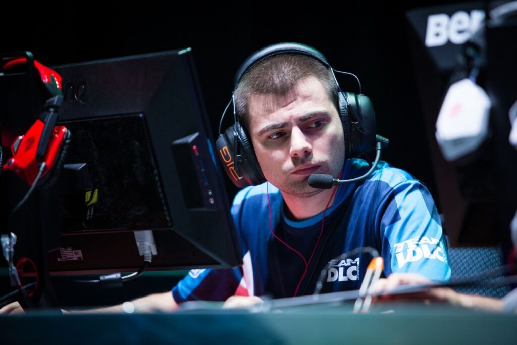 A close-up photograph of professional Counter-Strike player Hovik "KQLY" Tovmassian sitting at a desk during a competition, wearing headphones and a blue jersey with "TEAM DLC" printed on it