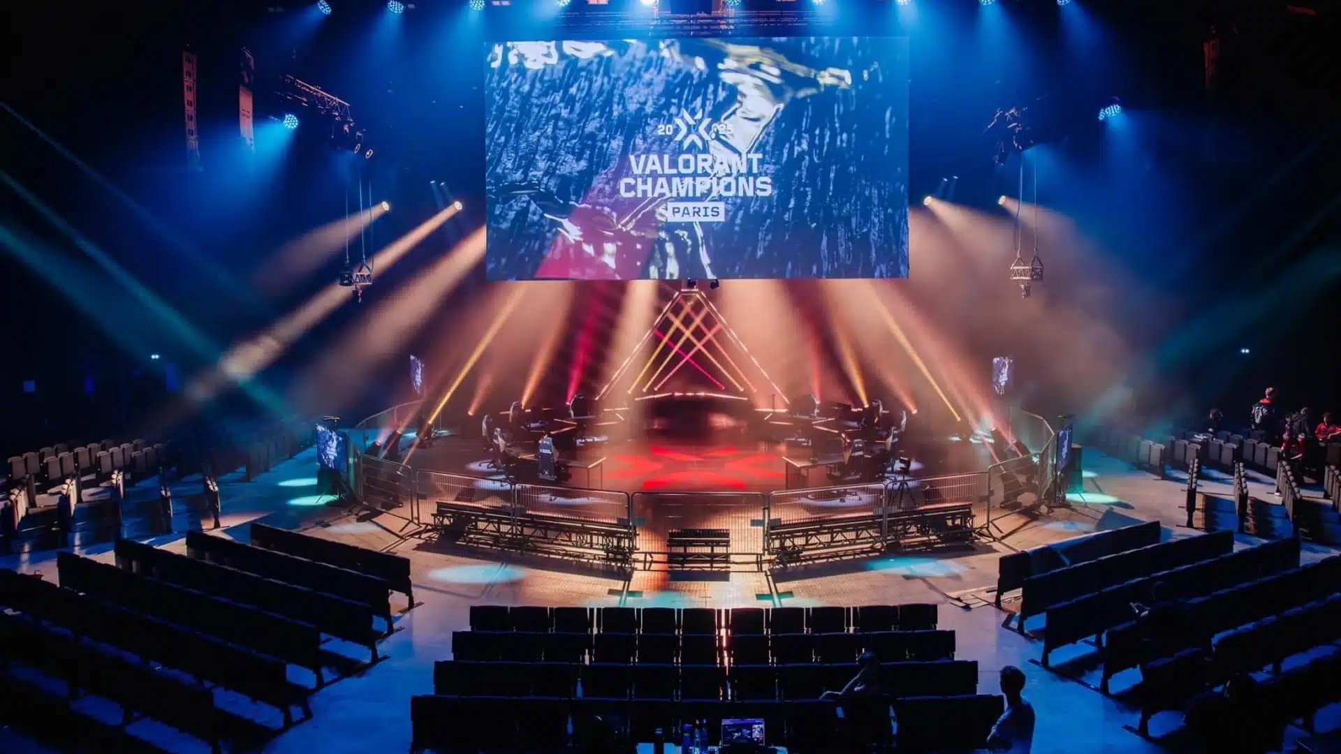 An esports stage for VALORANT Champions Paris with a large screen, red and blue lighting, and empty seating.