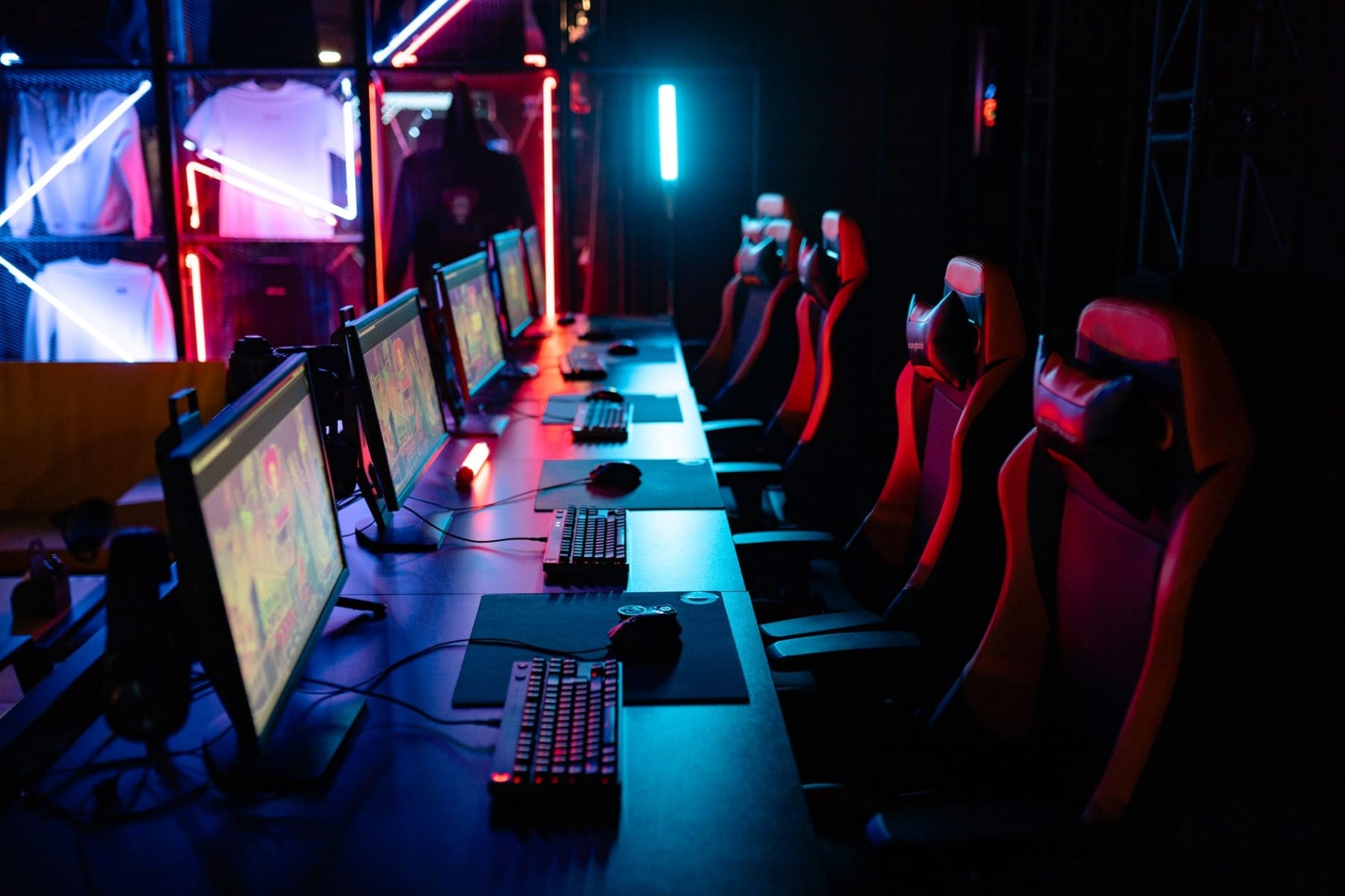 Row of gaming PCs, keyboards and red gaming chairs under neon lighting